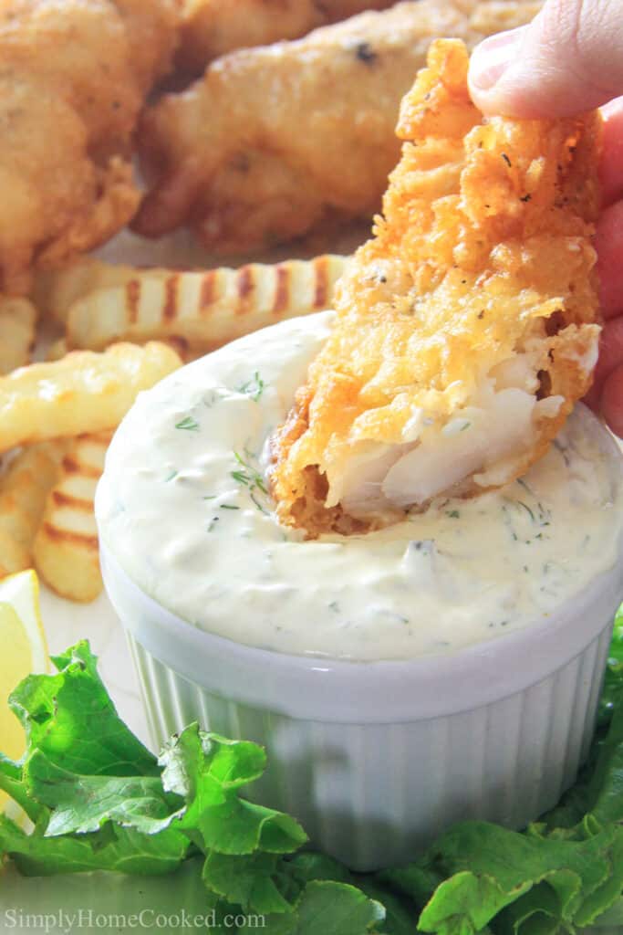 Beer Battered Cod is an amazing comfort food recipe perfect for gatherings with friends. Follow my easy recipe to make a batch of this crispy flavorful recipe! #fishandchips #beerbatteredcod #friedfrish #crispyfish #friedcod #easycod Beer Battered Cod being dipped in tartar sauce with fries in the background.