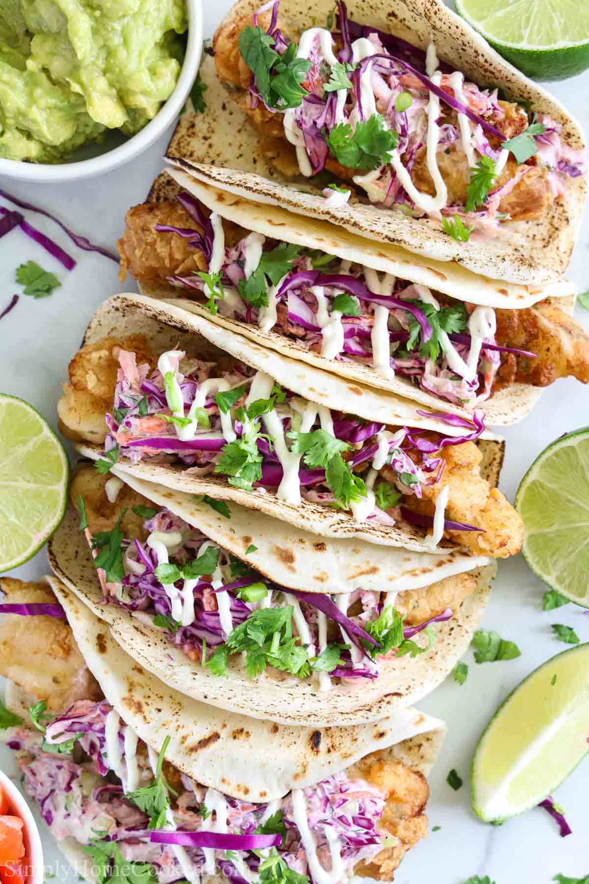 Irresistibly crispy fish tacos with baja cream and cabbage slaw. This is literally a flavor explosion! Crunchy, creamy, and bursting with delicious flavor! #fishtacos #bajatacos #bajacreamsauce #baja #crispyfishtacos Row of Fish Tacos with limes and guacamole on the side