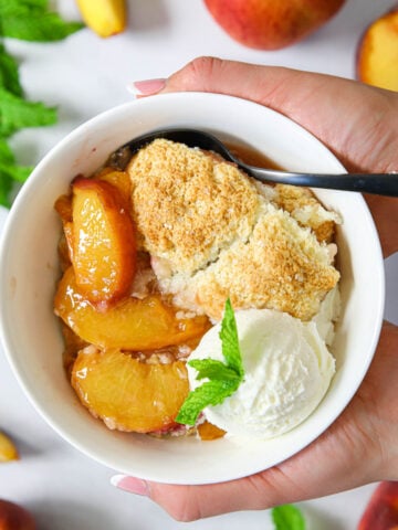 Hands holding a bowl of Easy Peach Cobbler with ice cream and a spoon.