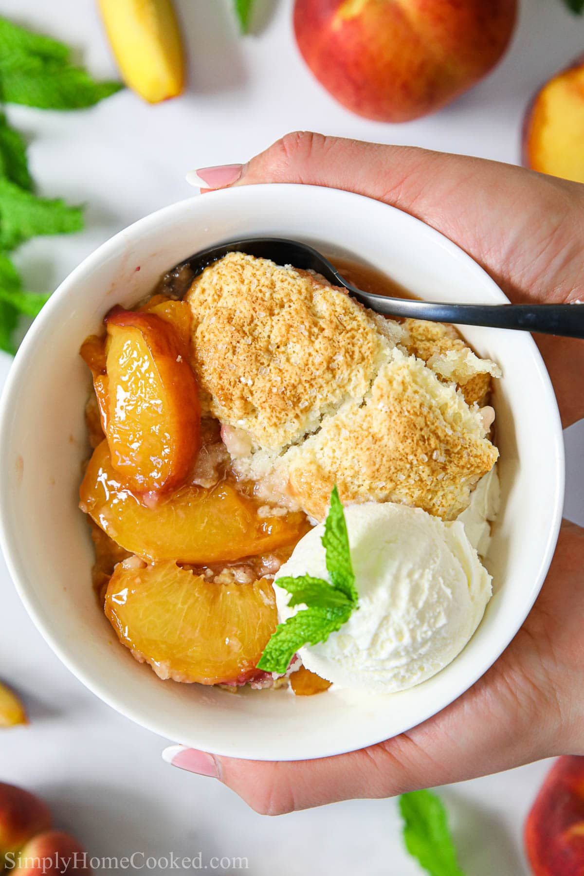 This easy homemade peach cobbler recipe will become your favorite summer dessert. Juicy peaches topped with a buttermilk biscuit topping and baked to perfection! #peachcobbler #peachcrisp #peachpie #peachdessert #summerdessert #cobbler Hands holding a bowl of Easy Peach Cobbler with ice cream and a spoon.