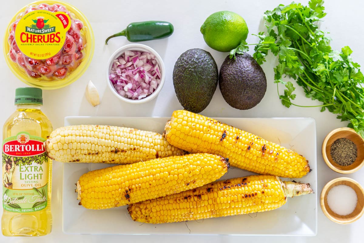 Ingredients for Avocado Corn Salad, including corn, avocado, tomatoes, cilantro, red onion, garlic, lime, jalapeno, olive oil, salt, and pepper.