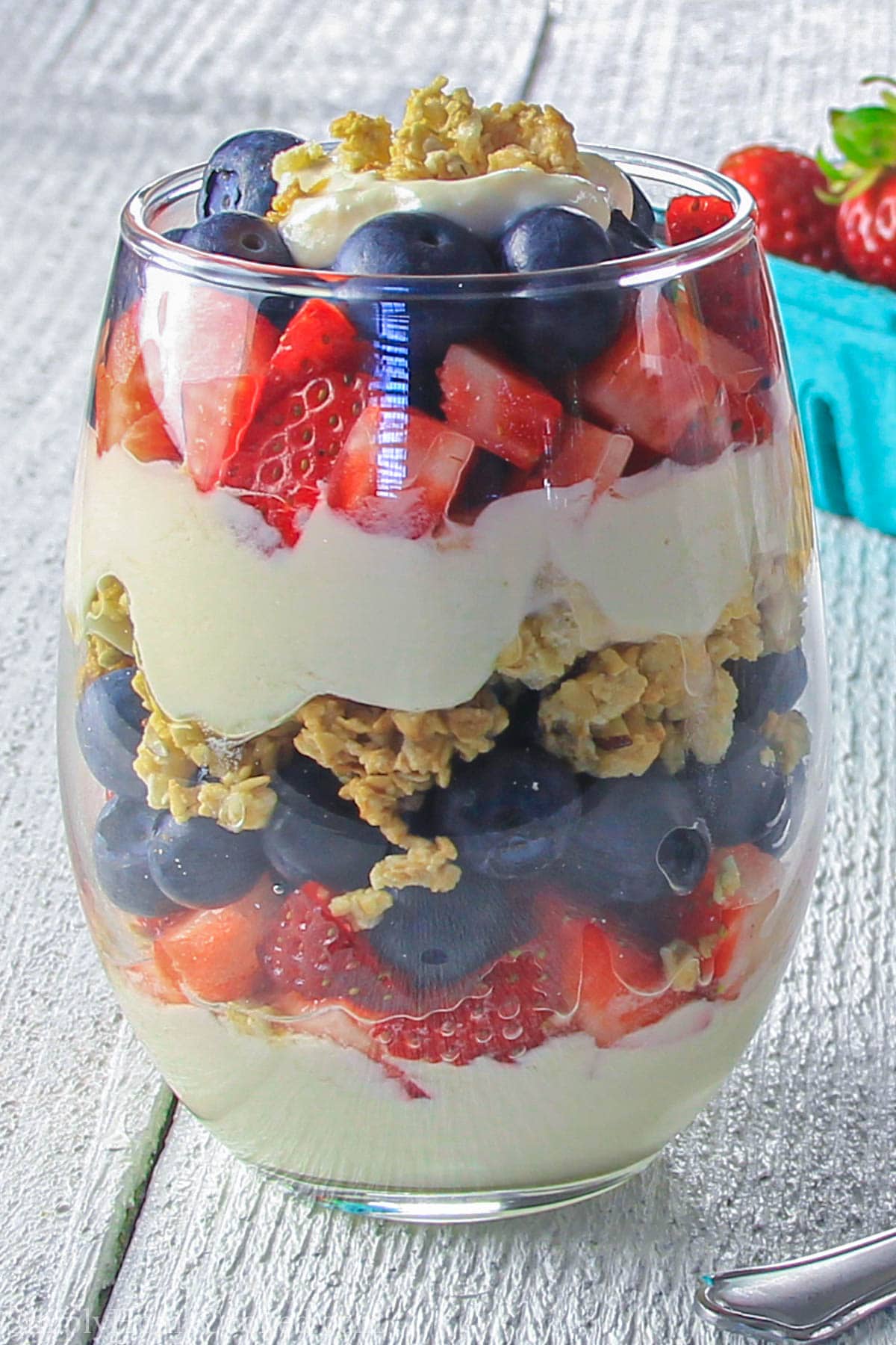This Greek yogurt berry parfait is loaded with blueberries, strawberries, honey vanilla Greek yogurt, and crunchy granola. #parfait #berryparfait #yogurtparfait #parfaitrecipe #fruitparfait Vertical image of a Yogurt Parfait in a glass, with strawberries in the background.