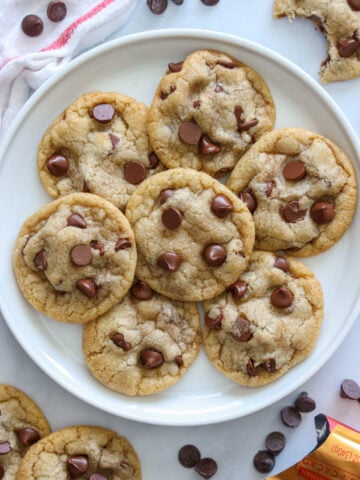 Vertical image of a plate of Chewy Chocolate Chip Cookies with cookies and chocolate chips scattered nearby.