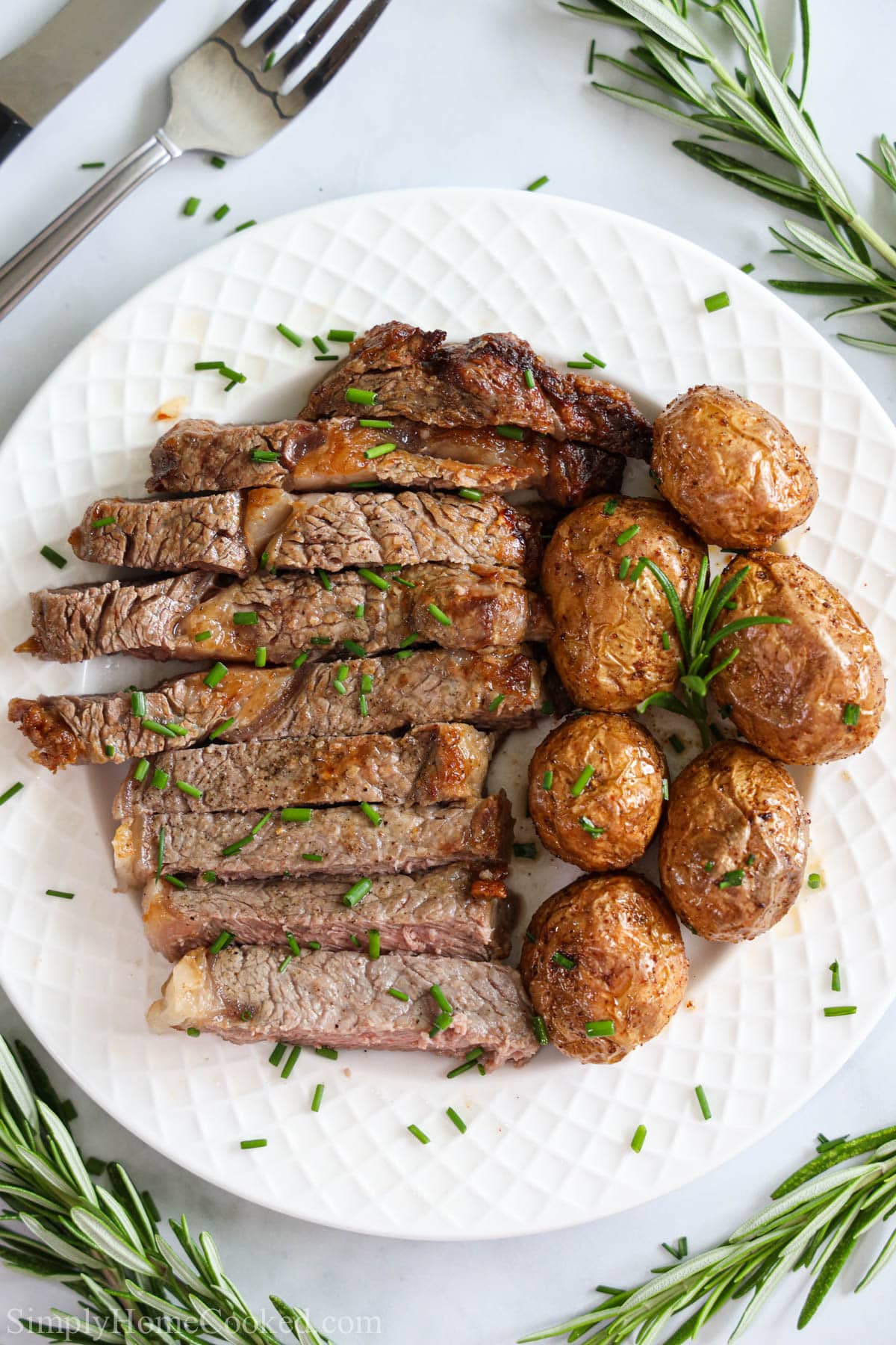 Vertical image of Juicy Air Fryer Steak sliced with potatoes on a white plate, rosemary sprigs nearby