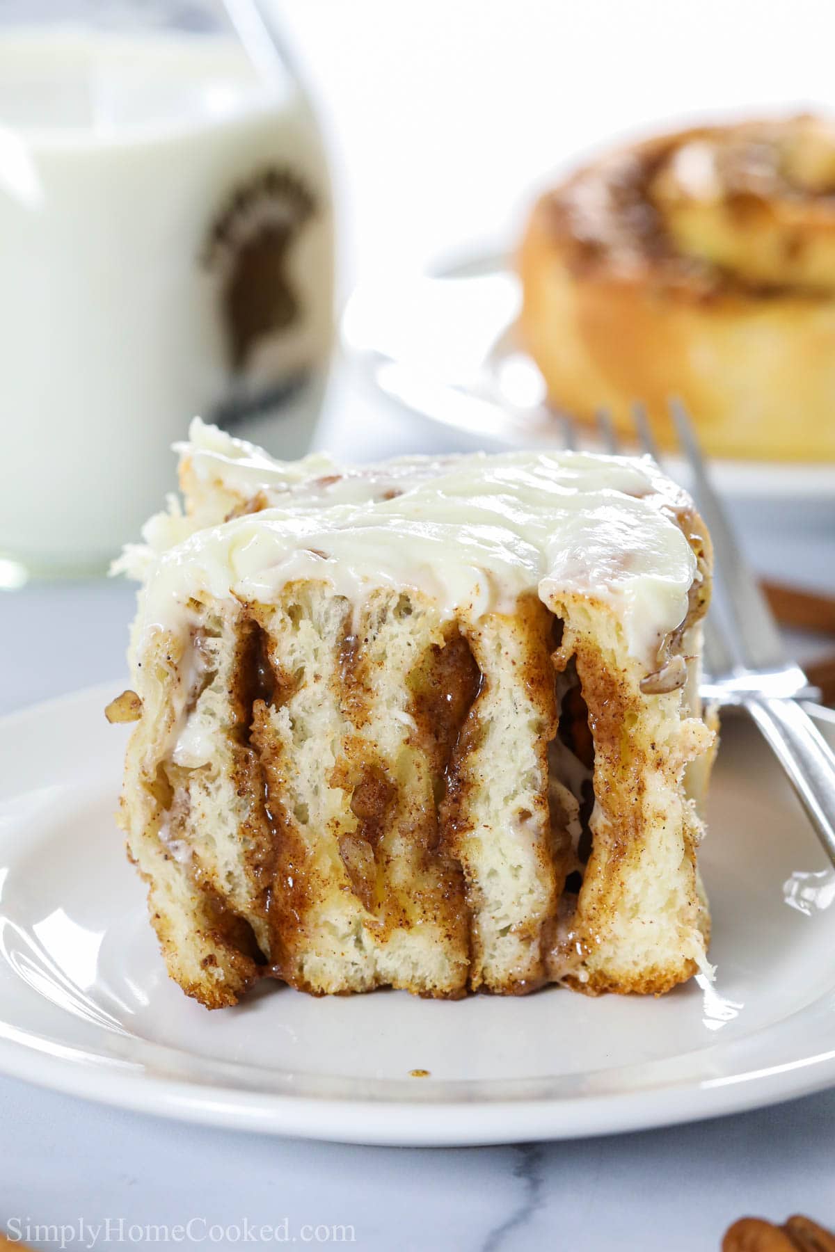 Delicious and fluffy Homemade Cinnamon Rolls topped with a cream cheese frosting, then filled with a cinnamon, brown sugar, and pecan filling. The dough can be made the night before so its an overnight recipe too! #cinnamonrolls #homemadecinnamonrolls #cinnaboncopycat #stickybuns Vertical image of Best Homemade Cinnamon Roll on a white plate with a fork, covered in cream cheese icing
