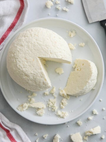 Vertical image of a plate of Farmer's Cheese molded with a slice cut out and crumbles nearby