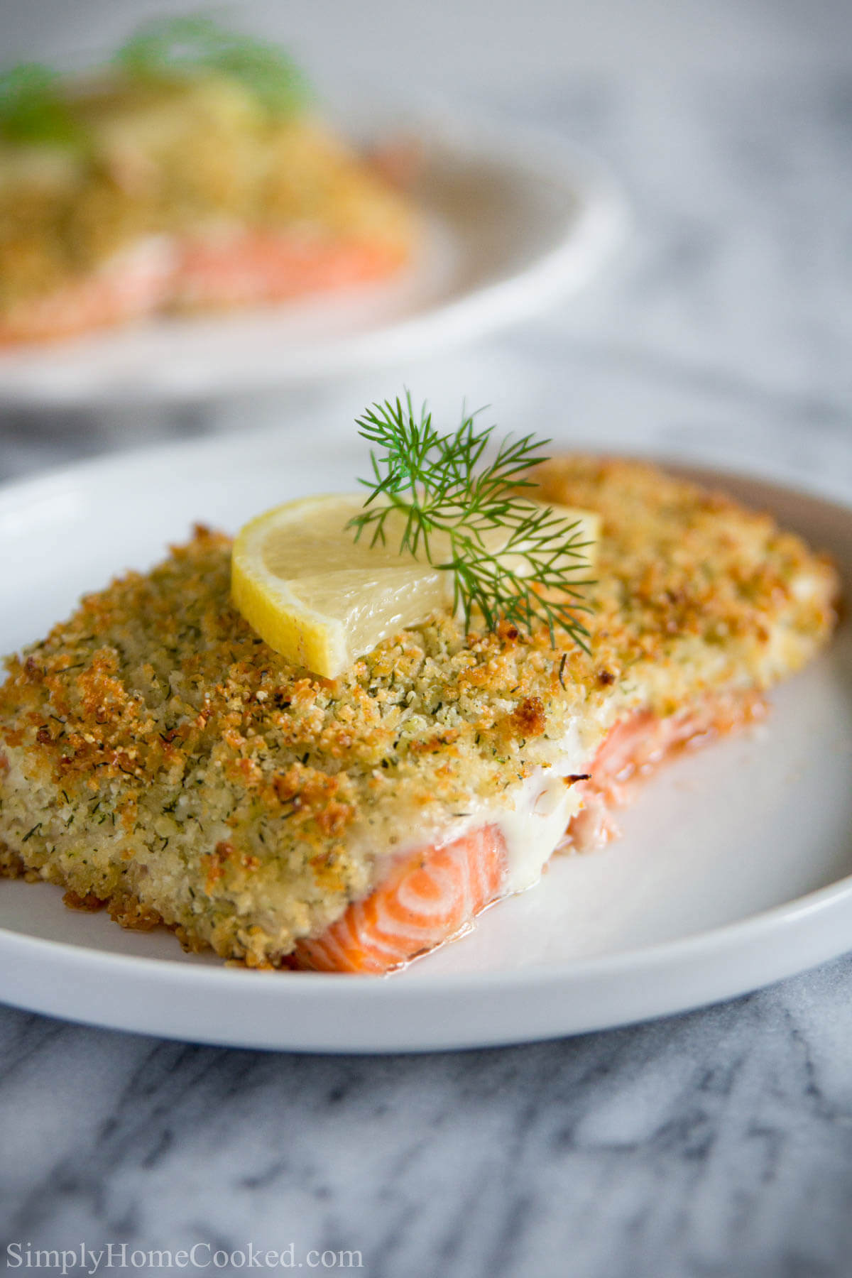 Vertical image of a plate of Panko Crusted Salmon topped with dill and lemon, another plate in the background