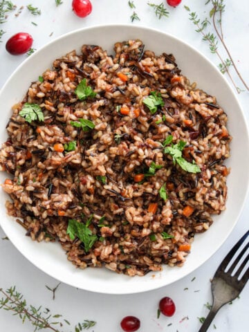 Vertical image of a bowl of Wild Rice with a fork, springs of thyme, and cranberries nearby