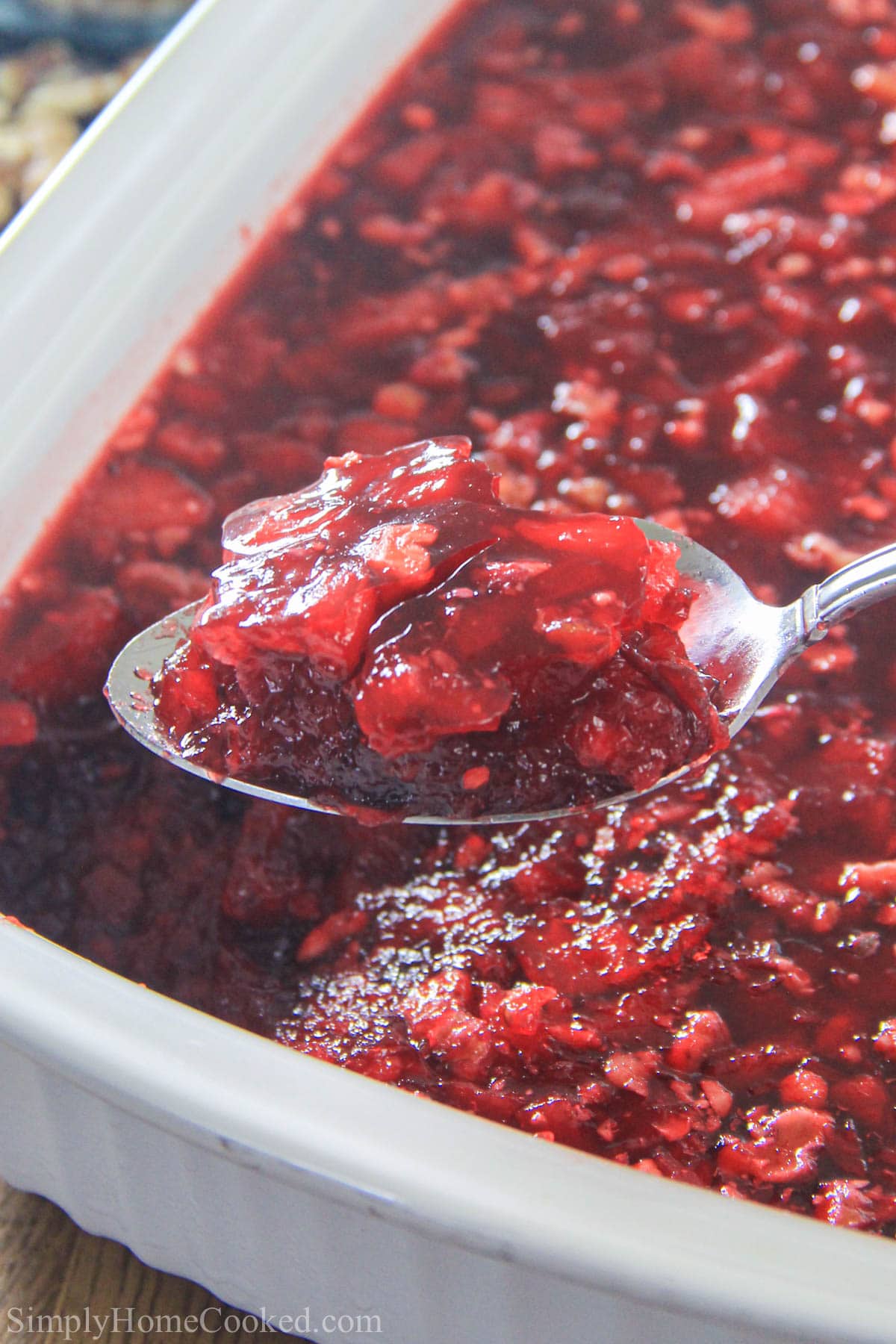 This Cranberry Jello Salad is always on our holiday table. The combination of tart and sweet go perfectly together. This recipe is a must for every table. A perfect side to your Thanksgiving turkey. #cranberryjello #cranberrysauce #thanksgivingsauce #thanksgivingfood #thanksgivingjello Vertical image of a spoonful of Cranberry Jello Salad lifted from a pan