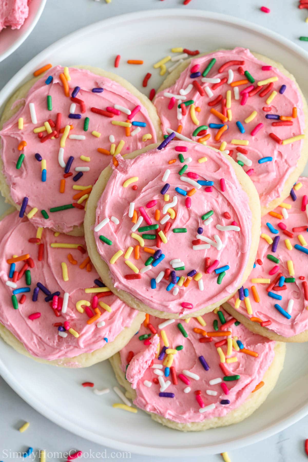 These Frosted Lofthouse Sugar Cookies are perfect for the holidays, or anytime. Soft, cake-y, and sweet, they practically melt in your mouth. #frostedcookies #frostedlofthousecookies #frostedsugarcookies #frostedlofthousesugarcookies overhead image of pink lofthouse cookies with rainbow sprinkles on top.