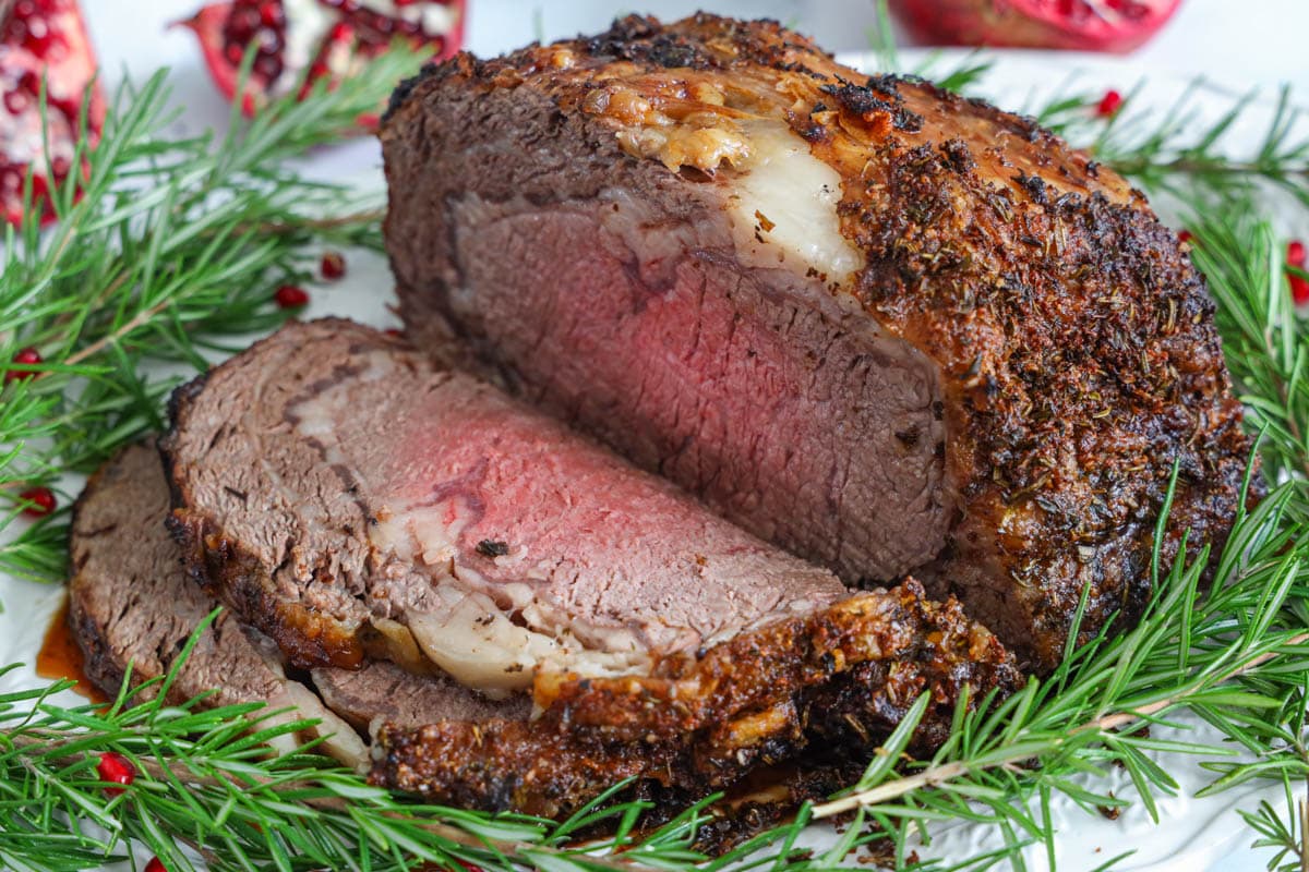 A seasoned herb crust and a juicy center, this prime rib roast will become a favorite in your home. #primerib #bestprimerib #easyprimeribdinner #homemaderibroast Prime Rib sliced on a plate with herbs and pomegranate seeds