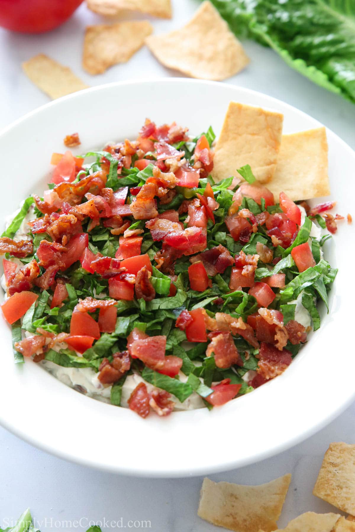 This Easy BLT Dip is creamy, savory, and delicious. Enjoy it with your favorite veggies or chips as a snack or serve it to guests as a tasty appetizer. #easybltdiprecipe #bestbltdiprecipe #homemadebltdip #bltdipapp Vertical overhead image of a bowl of Easy BLT Dip with pita crackers to the side