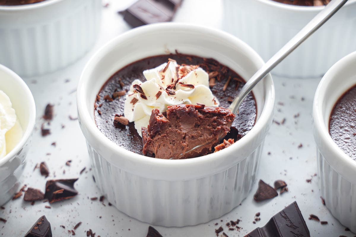 Chocolate Pot De Creme topped with whipped cream, a spoonful being lifted out, and chocolate shavings nearby
