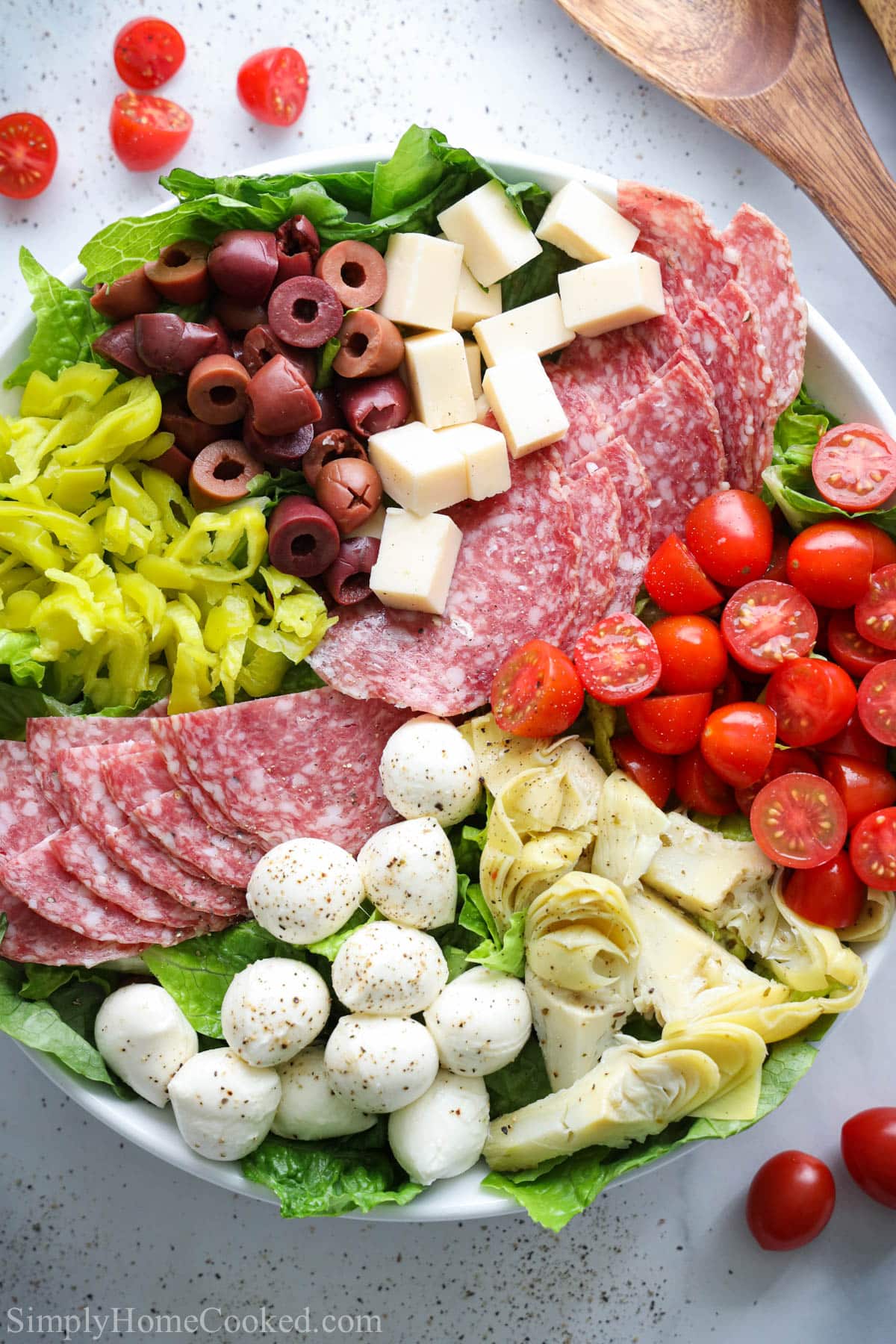 This Antipasto Salad is a delicious way to start your meal. Full of salami, sun-dried tomatoes, olives, and other veggies, it makes a great appetizer. #Italianappetizer #antipastosalad #antipastosaladrecipe #antipastorecipe image of Antipasto Salad in a white plate with a wooden spoon next to it