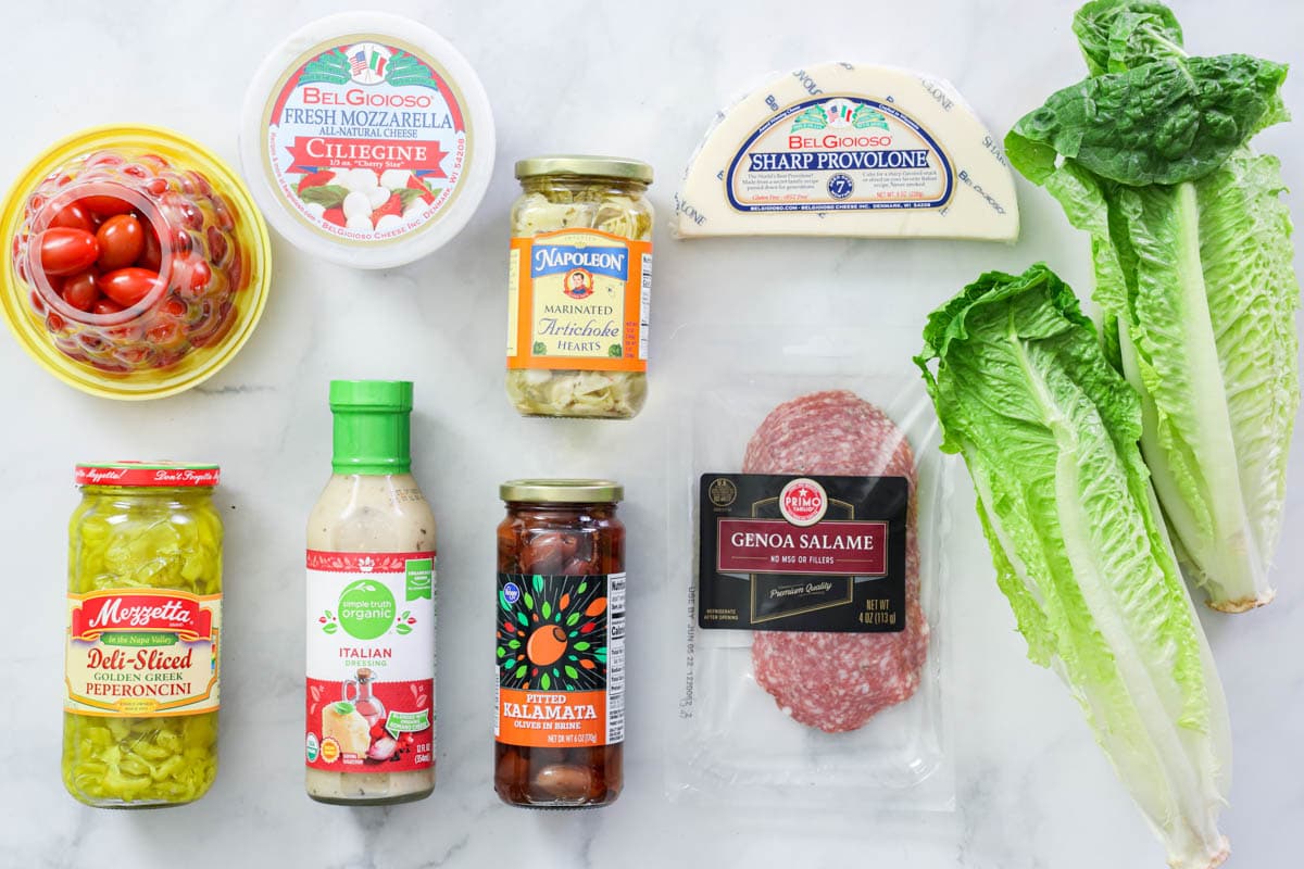 Ingredients for Antipasto Salad, including lettuce, tomatoes, mozzarella, provolone, salami, lives, artichoke hearts, pepperoncini, and Italian dressing.