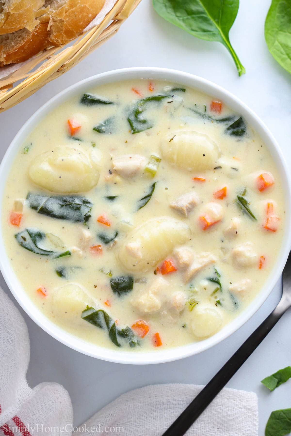 Vertical overhead image of a bowl of Chicken Gnocchi Soup with basket of bread, spinach, and a spoon nearby 