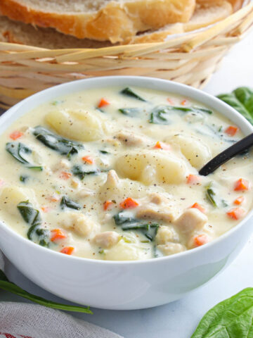 Vertical image of a bowl of Chicken Gnocchi Soup with basket of bread nearby and a spoon in it.