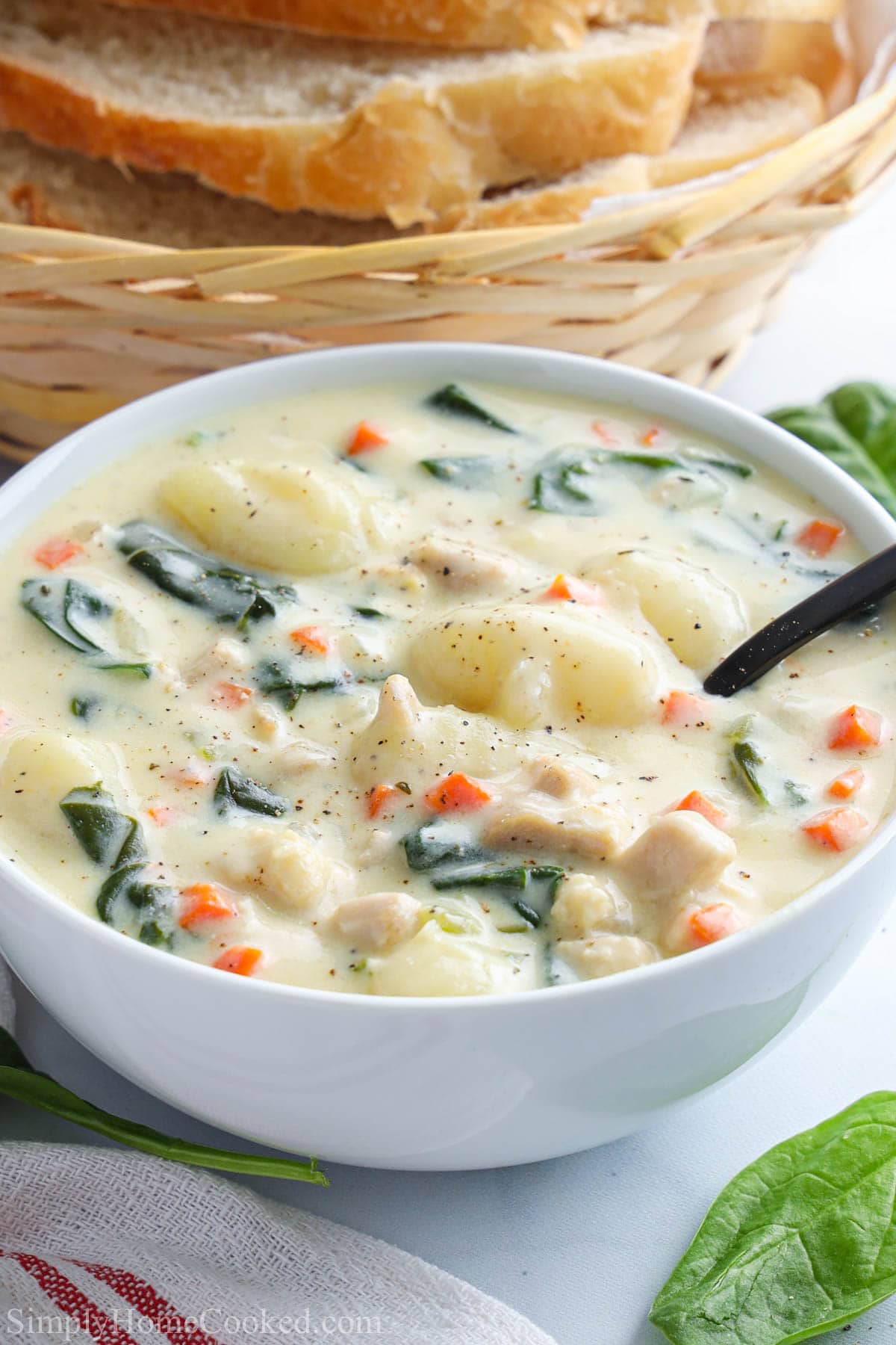 A bowl of Chicken Gnocchi Soup with basket of bread nearby and a spoon in it.