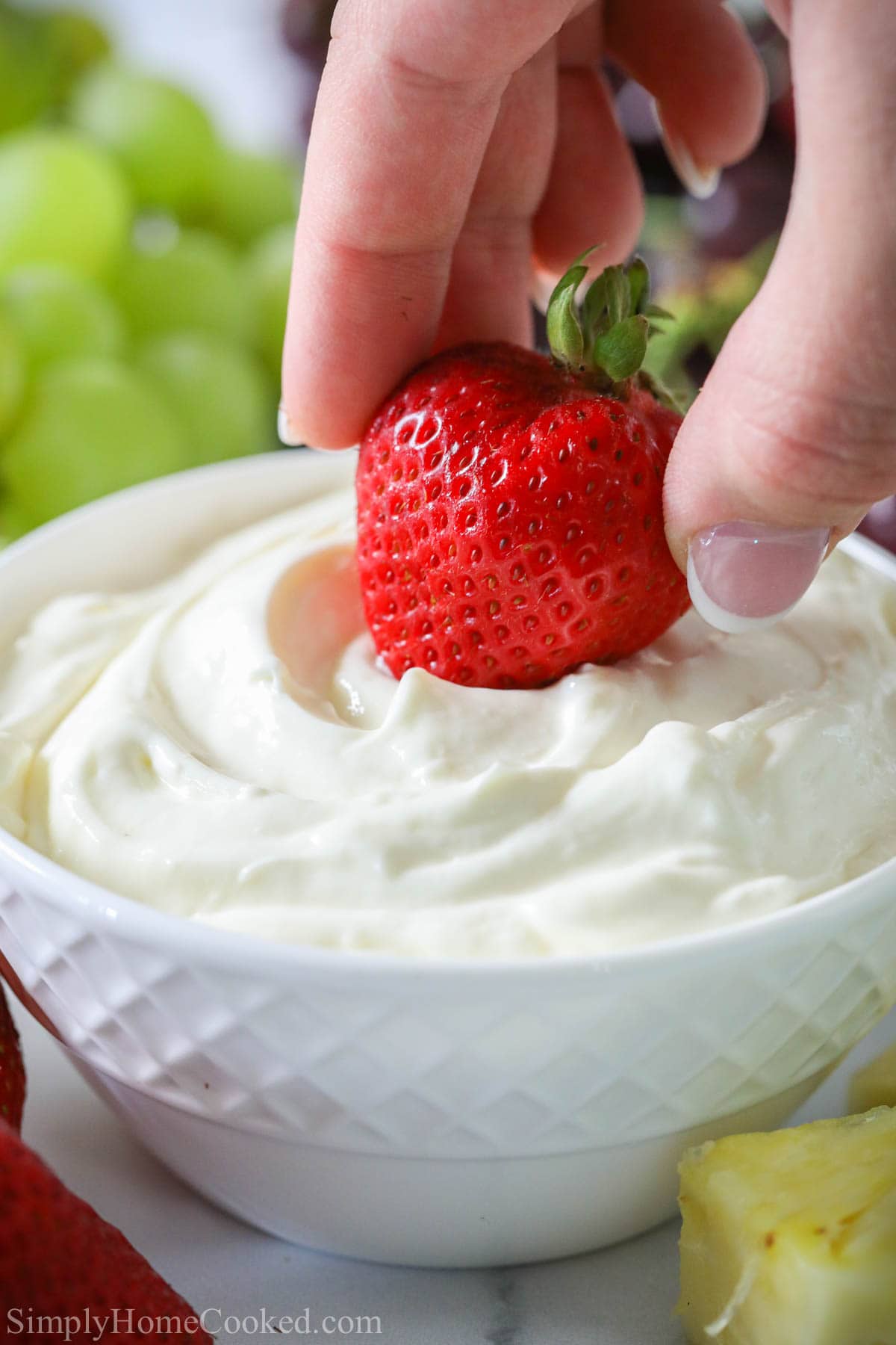 This easy cream cheese fruit dip recipe will become your favorite dip for almost any fruit. With only 4 ingredients, it comes together so quickly! #fruitdip #creamcheesedip #berriesandcream #easyfruitdip #fruitplatter Hand dipping a strawberry into Cream Cheese Fruit Dip in a white bowl