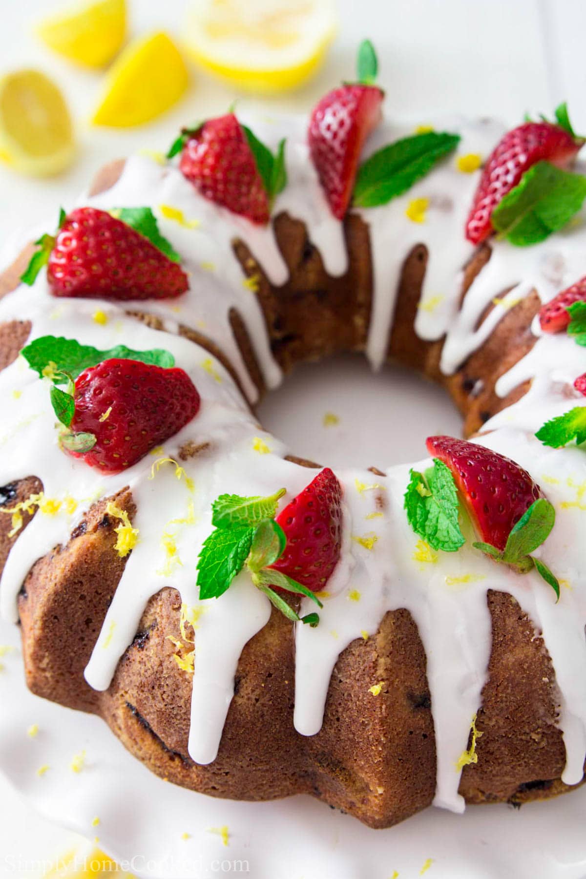 Make this Homemade Strawberry Bundt Cake Recipe as a beautiful and delicious treat everyone at the table will love. This easy recipe is loved by all! #bundtcake #strawberrycake #summercake #berrycake #strawberrybundt Strawberry Bundt Cake with glaze and strawberries on top with mint leaves and lemons nearby
