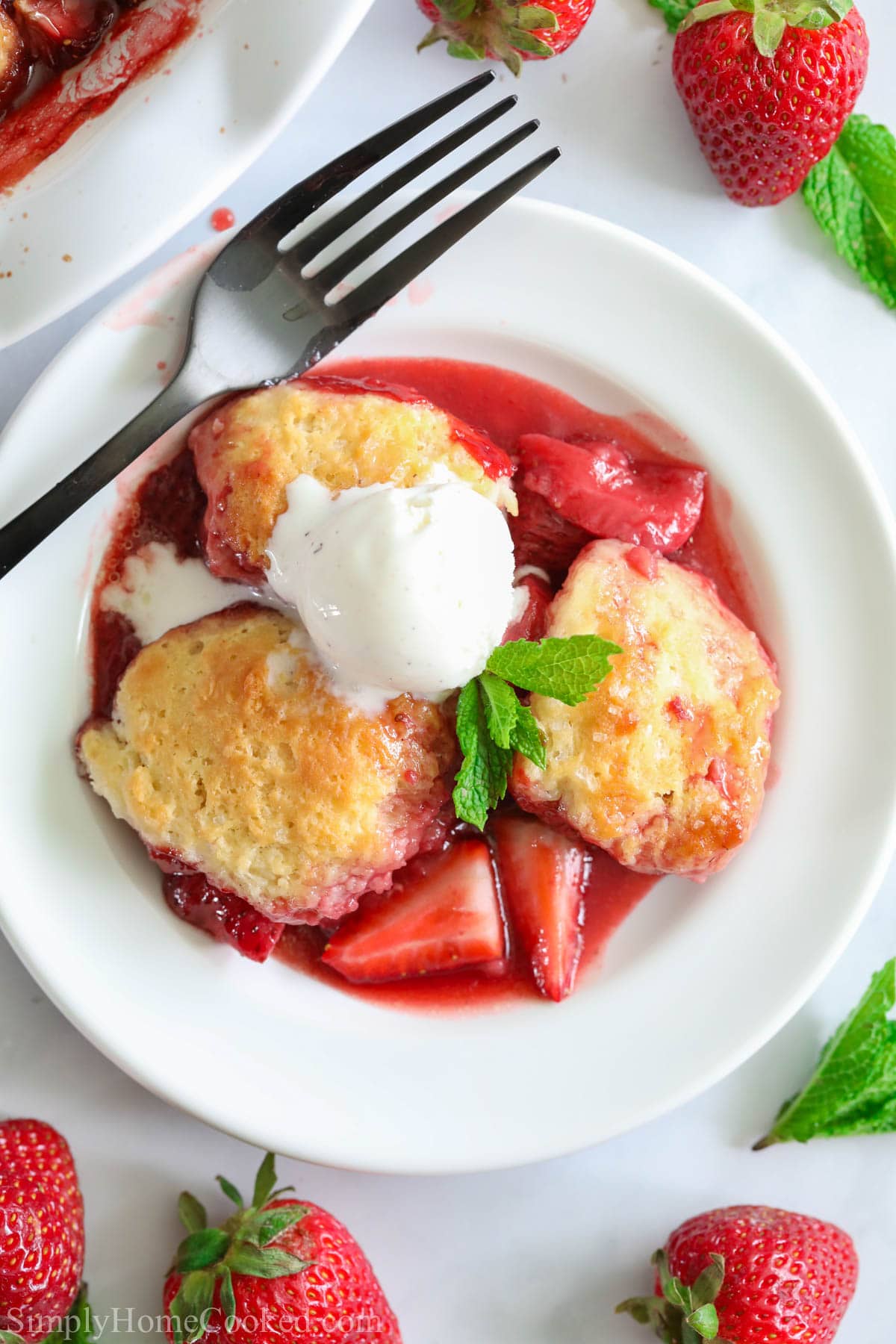 This Strawberry Cobbler is simple & sweet. With ripe berries & a golden biscuit crust, it's a delicious dessert that's the perfect end to a summer night. #easycobblerrecipe #easyhomemadecobbler #easyhomemadestrawberrycobbler #bestcobbler Strawberry Cobbler with ice cream on a white plate with a fork and strawberries nearby