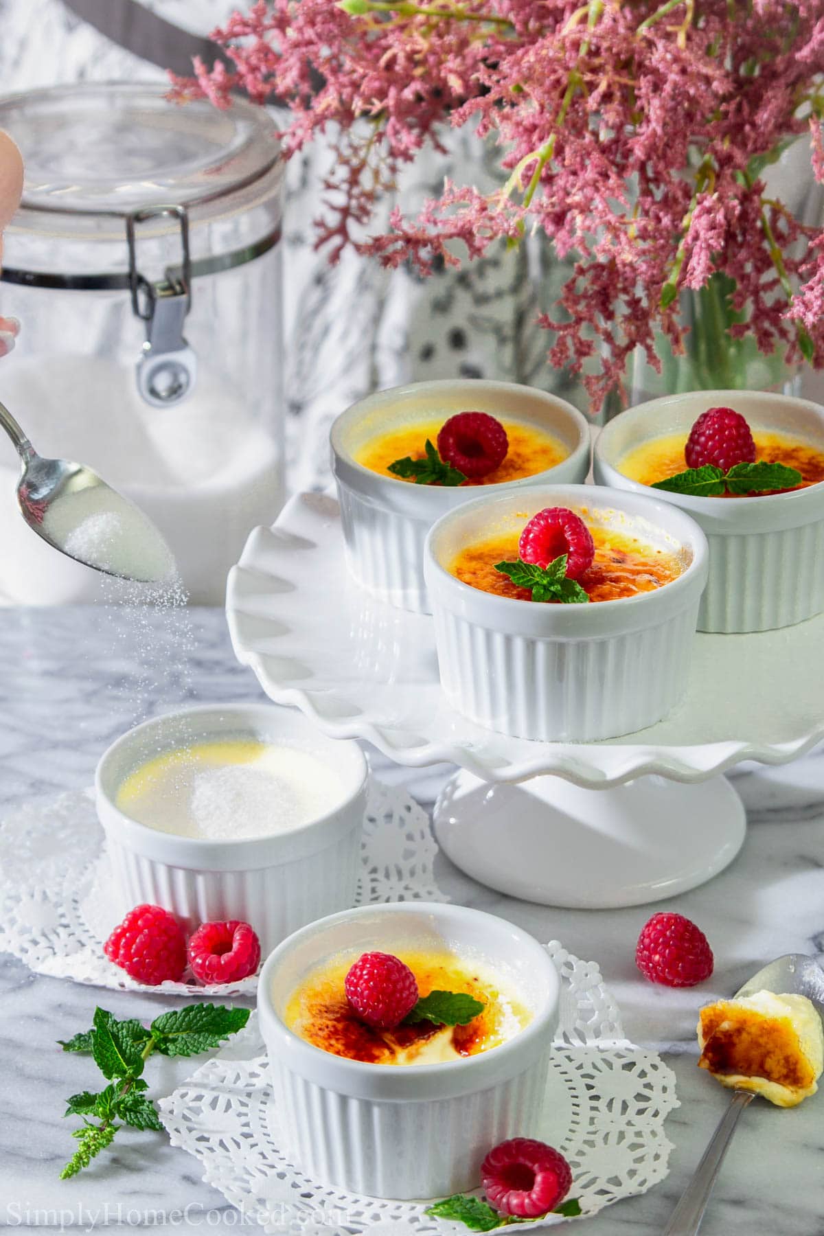 Ramekins of White Chocolate Creme Brulee on a lazy susan, and more on the table, topped with fresh mint and berries, as well as sugar from a spoon.