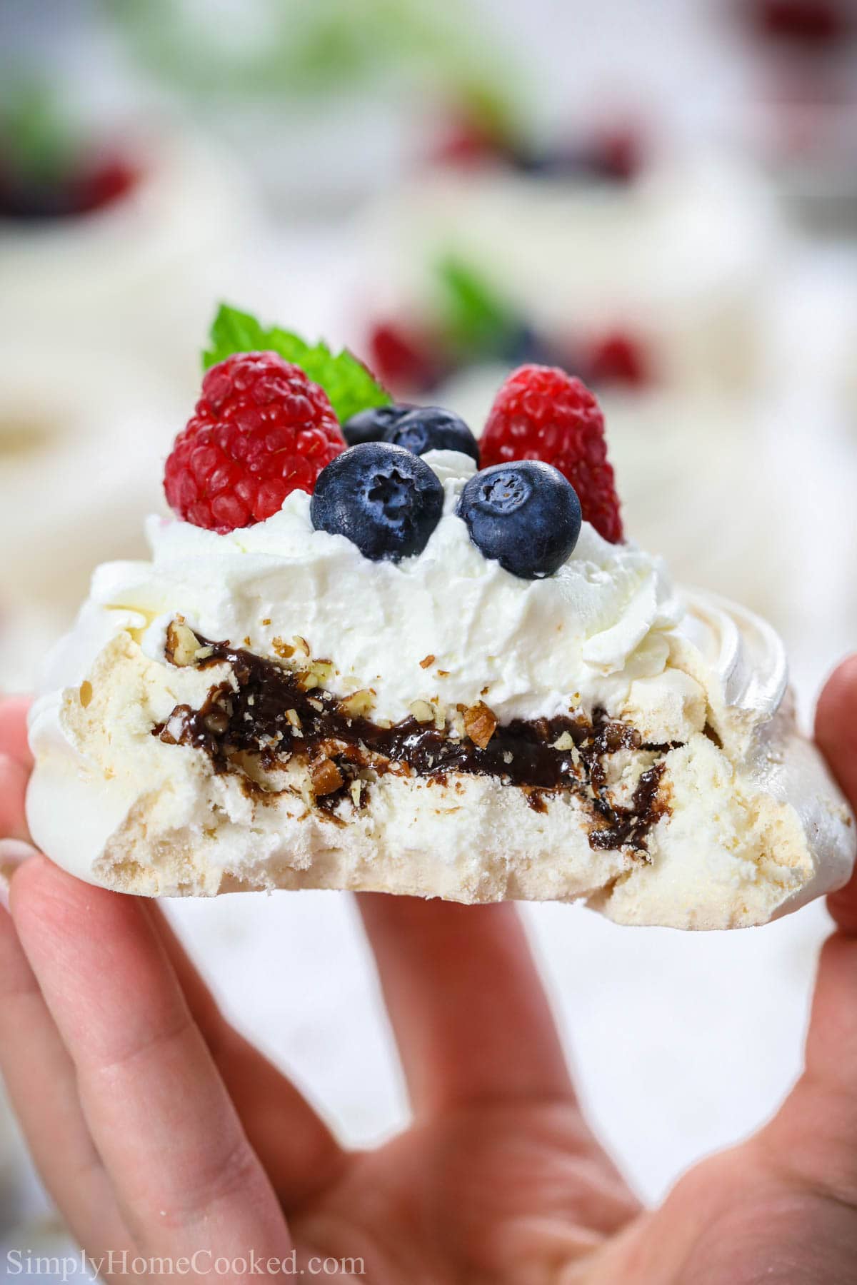 Mini crispy meringue pavlova filled with chocolate ganache, chopped pecans, a simple whipped cream topping, and fresh berries. Best summer dessert recipe! #minipavlova #pavlova #meringue #summerdessert #pavlovarecipe Mini Pavlova with Chocolate and pecans and topped with whipped cream and berries, missing a bite and being held in a hand