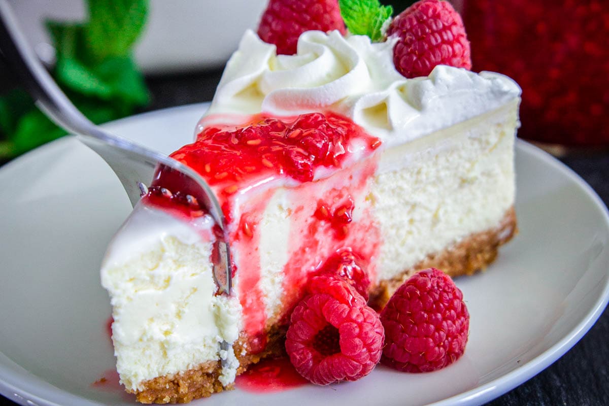 This classic New York style cheesecake recipe is rich, creamy, and insanely EASY to make. Learn all the secrets to a perfect smooth cheesecake without any water bath and no cracks! #newyorkcheesecake #classiccheesecake #cheesecake #whitecheesecake #vanillacheesecake Slice of New York Style Cheesecake on a white plate and topped with raspberry sauce and fresh berries, a fork breaking off a piece.