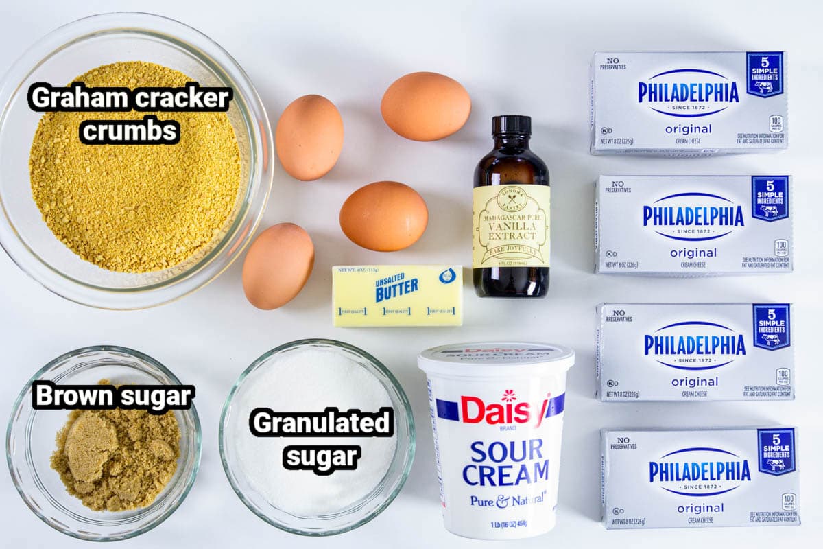 Ingredients for New York Style Cheesecake, including graham crackers, brown sugar, granulated sugar, eggs, butter, vanilla, sour cream, and cream cheese.