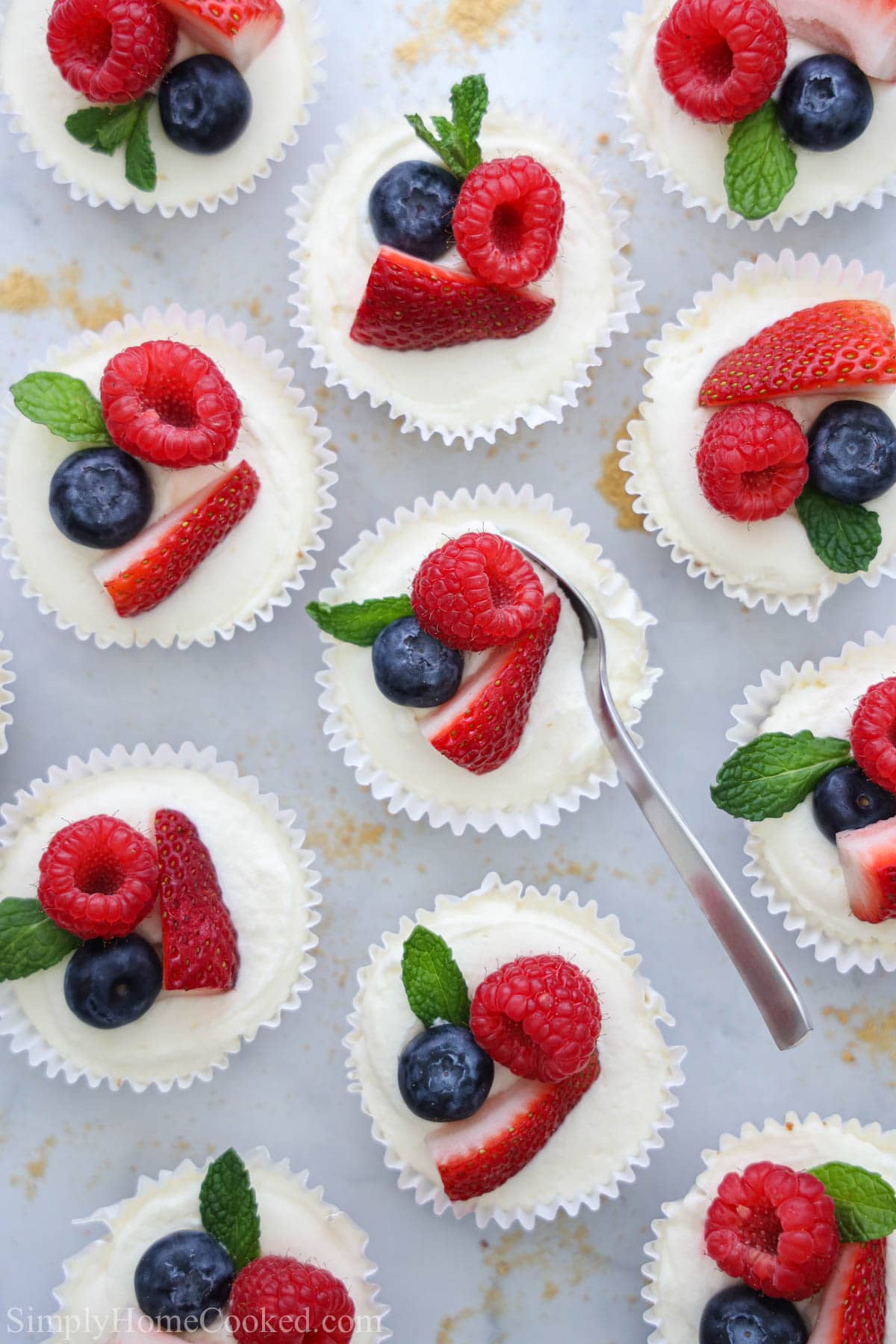 These No Bake Mini Cheesecakes are creamy, tangy, and delicious. Easy to make and richly flavored, these individual desserts are perfect for sharing. #nobakecheesecake #nobakeminicheesecake #minicheesecake #nobakeminicheesecakes Overhead of No Bake Mini Cheesecakes topped with fresh berries and a spoon scooping into one of them