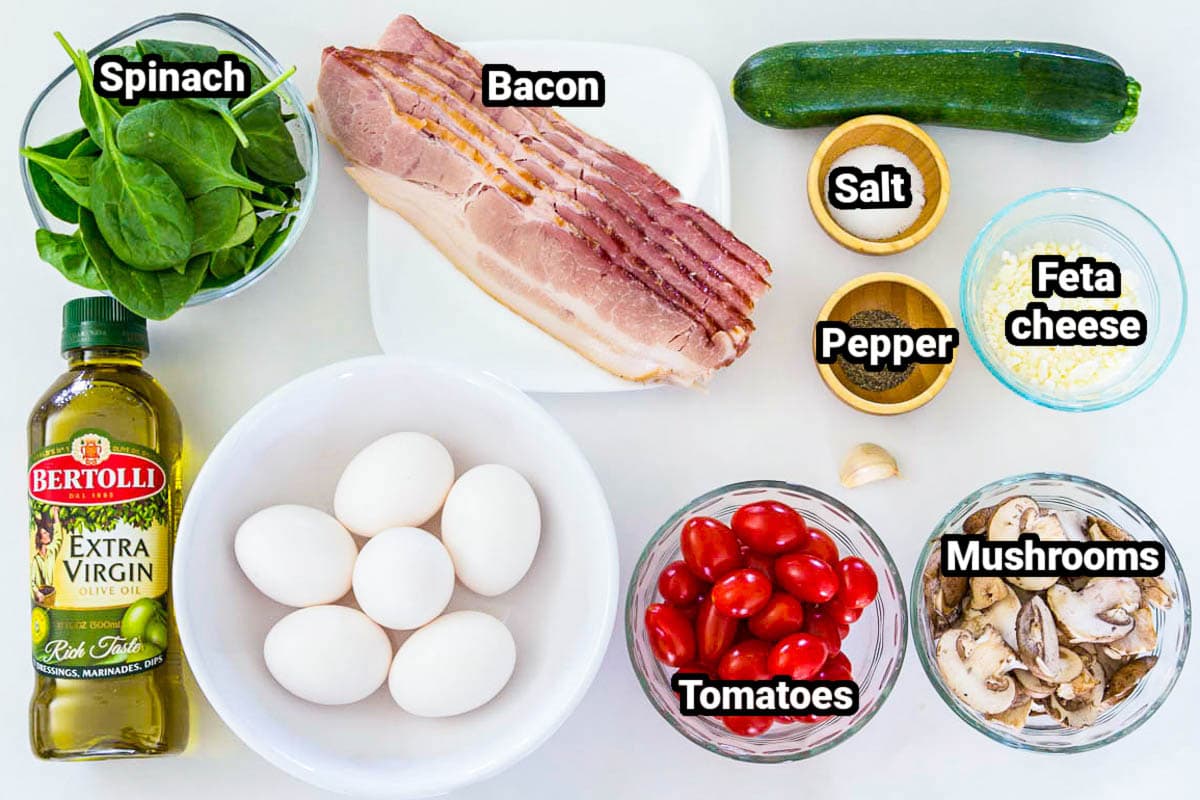 Ingredients for Zucchini Frittata, including spinach, olive oil, bacon, eggs, salt, pepper, zucchini, tomatoes, mushrooms, garlic, and feta cheese.
