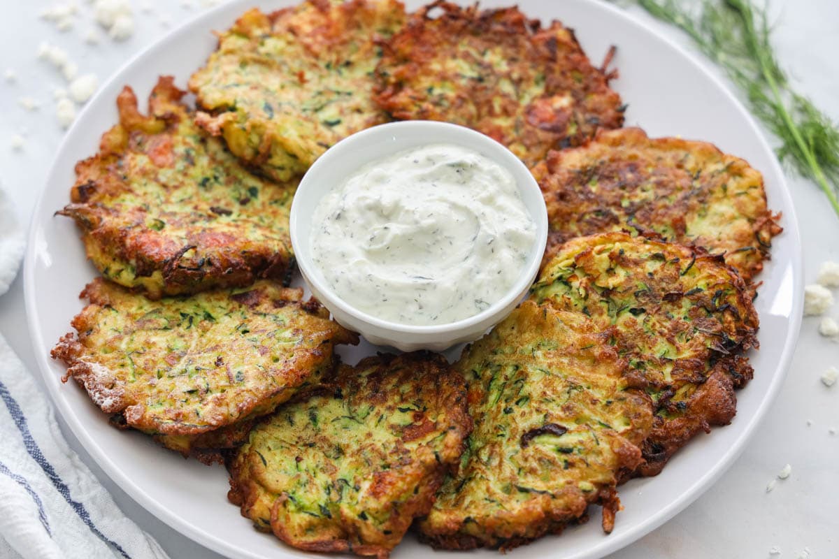 Zucchini Fritters circled on a white plate with tzatziki sauce in the middle