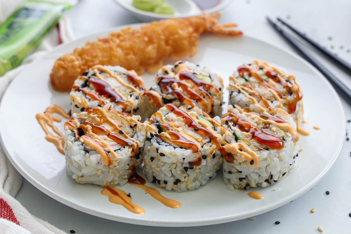 Sliced Dynamite Roll with unagi sauce and spicy mayo drizzled on top on a white plate with shrimp tempura