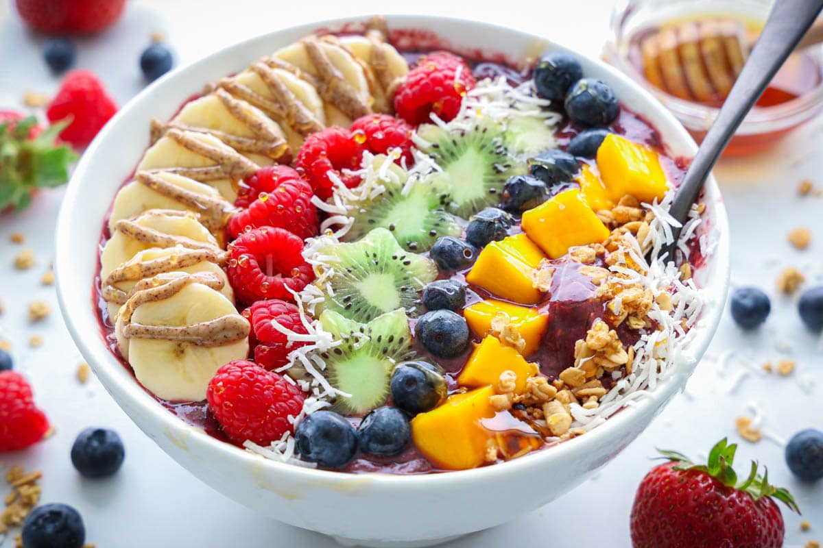 Acai Bowl topped with fresh fruit, coconut flakes, granola, honey, and nut butter, with strawberries and honey nearby and a spoon in the bowl.