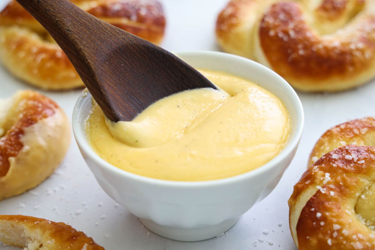 A wooden spoon stirring Pretzel Cheese Dip in a white bowl near soft pretzels.