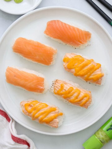 Salmon Nigiri on a white plate, some with spicy mayo on top, chopsticks and wasabi paste nearby