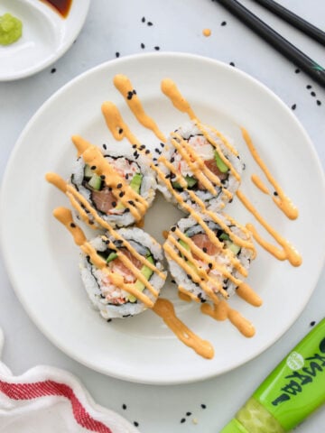 Spicy Salmon Roll cut up on a white plate with spicy mayo drizzled on top and chopsticks and wasabi nearby.