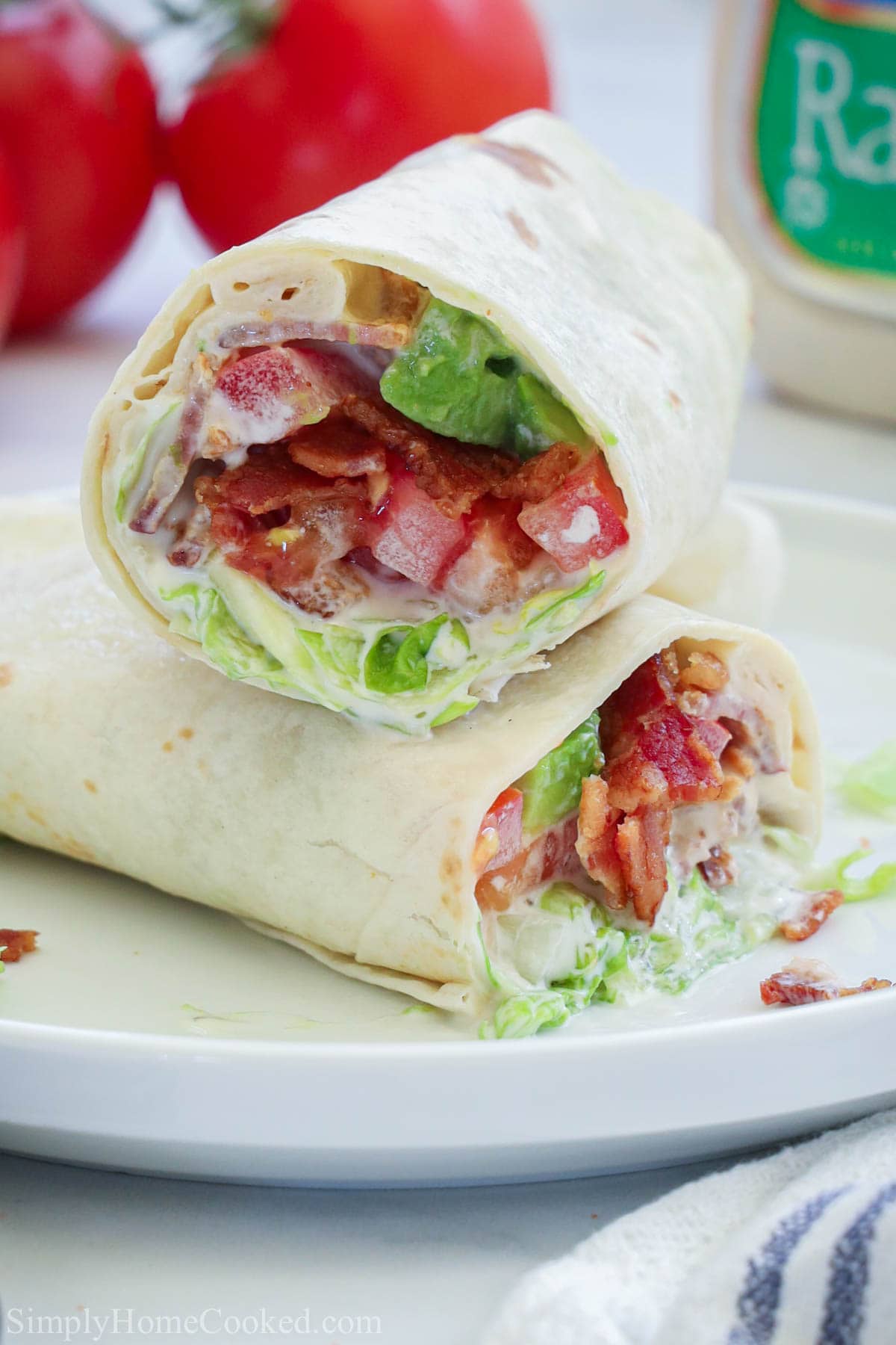 This easy to make BLT Wrap is the perfect on-the-go snack. You can even add it to your work lunch or your kids' school lunches for a filling and flavorful meal. #easybltwrap #blt #bltwrap #schoollunchideas Two halves of an Easy BLT Wrap laying crosswise on top of each other on a white plate.