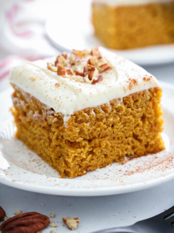 Slice of Pumpkin Cake on a white plate with pecans on top of the cream cheese frosting.