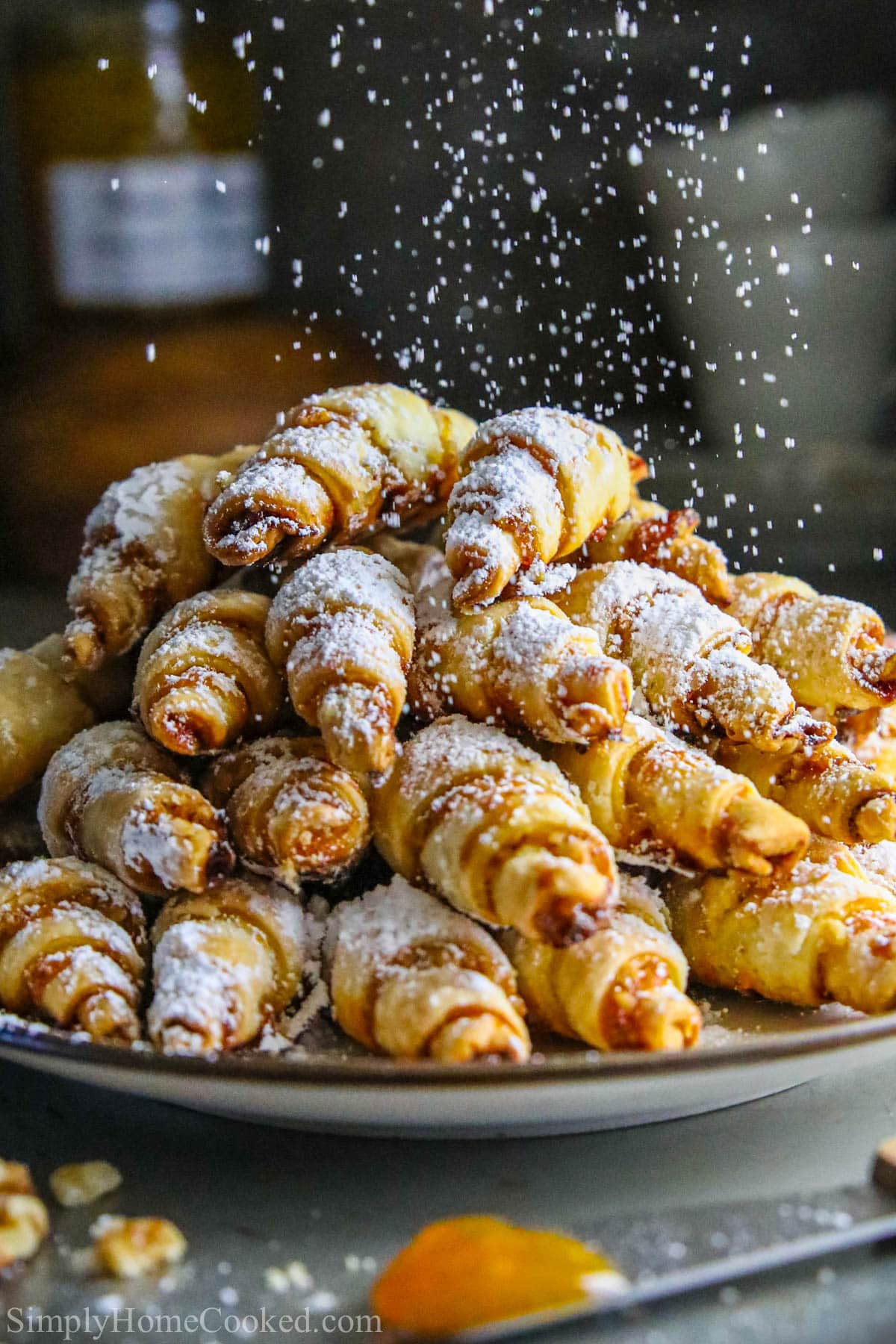 This is perhaps the easiest rugelach recipe you've ever seen! These apricot walnut rugelach cookies are filled with apricot jam and chopped walnuts. #rugelach #jewishcookies #christmascookies #holidaycookie #pastry #rogaliki Plate of Apricot Rugelach being sprinkled with powdered sugar.