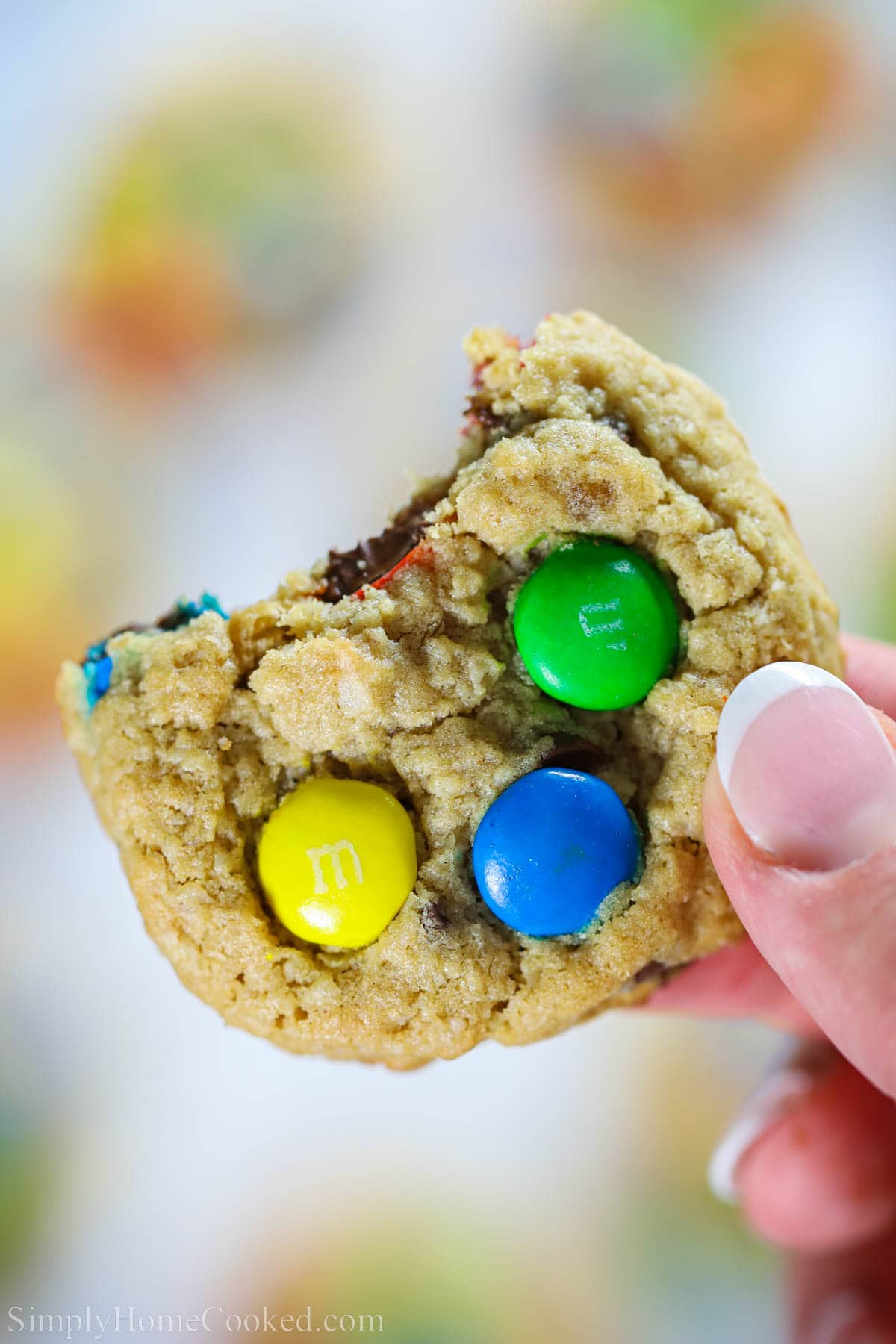 Delicious and soft, these thick Monster cookies will be a crowd pleaser. They are moist and chewy, with the perfect amount of chocolately goodness. #chocolatechipmonstercookiesrecipe #bestmonstercookie #cookierecipe #M&Mmonstercookies #chewyhomemadecookies Hand holding a Monster Cookie