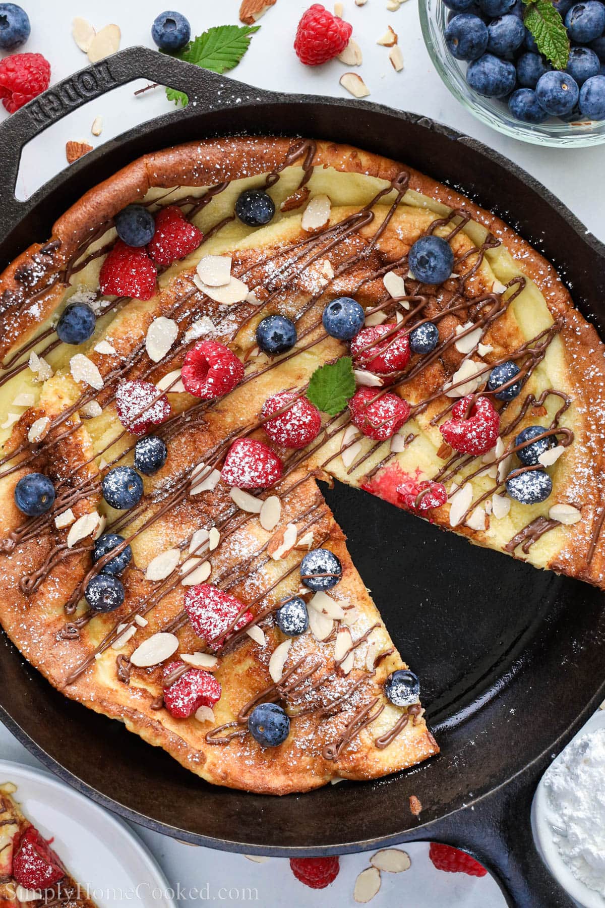 An easy and delciosu dutch baby pancake recipe that you will want to make time and time again. #dutchbaby #dutchbabypancake #germanpancake #nutellapancakes #onepanpancake German Pancake in a cast iron skillet topped with Nutella, sliced almonds, berries, and powdered sugar, one slice missing.