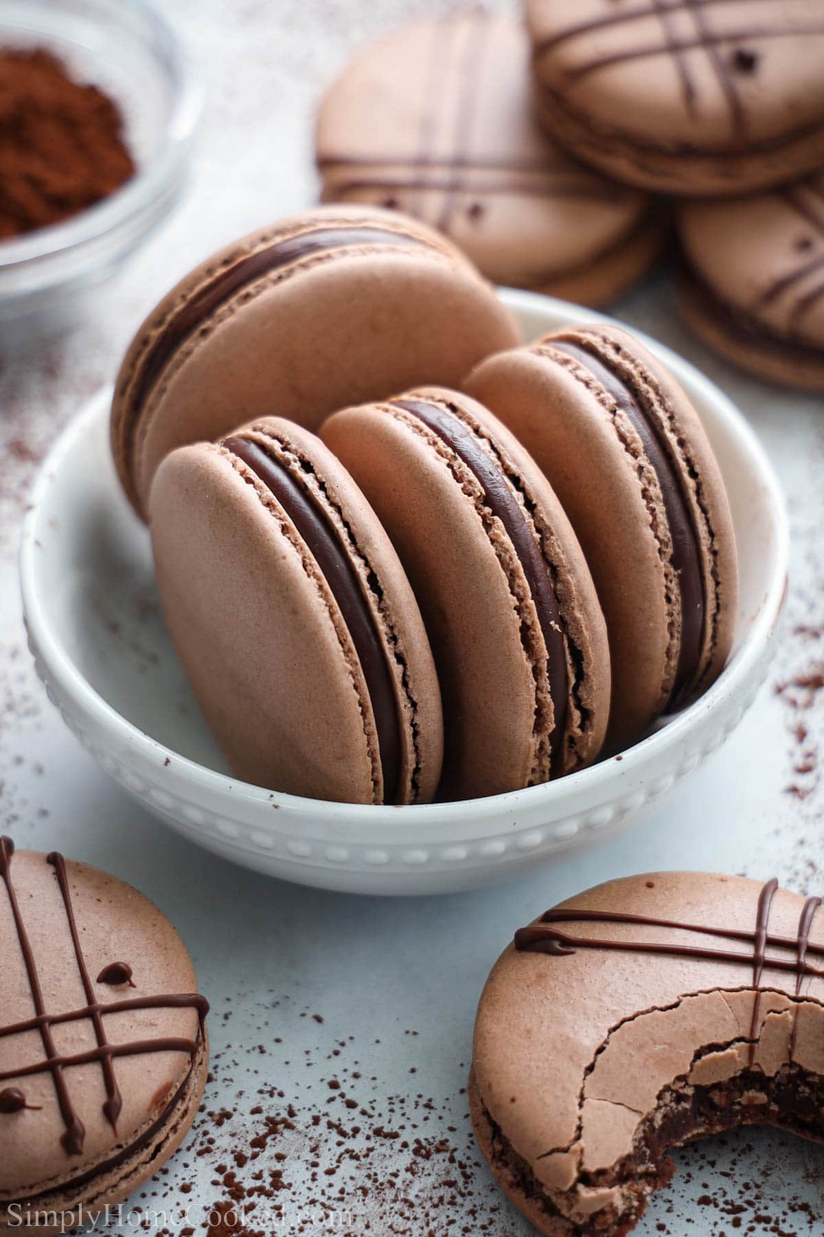 Chocolate Macarons with a ganache filling in a bowl with cocoa powder and macarons nearby.