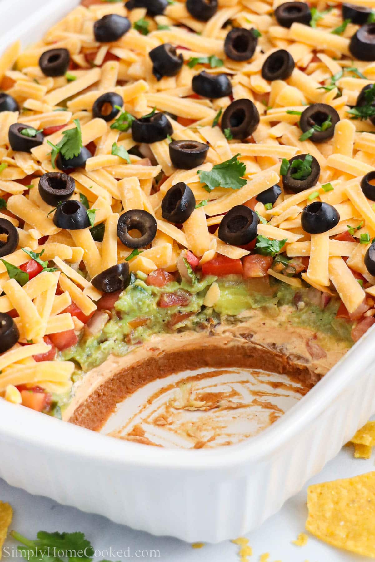 This 7 layer dip is layered with guacamole, pico de gallo, beans, sour cream, and olives. Its such a crowd pleaser! Perfect for parties or game day! #7layerdip #gamedayfood #mexicanappetizer #layereddip #diprecipe Baking dish of 7 Layer Taco Dip with a scoop missing.