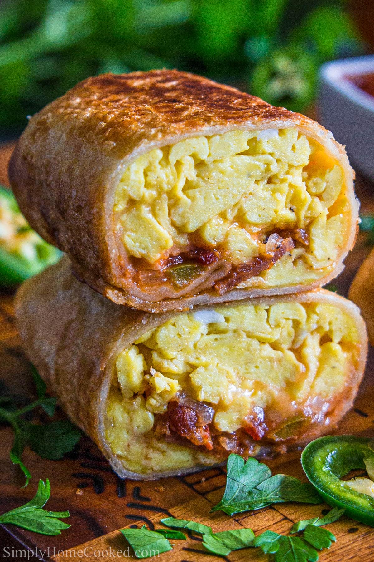 This Crispy Breakfast Burrito recipe will be a favorite in your family. Delicious eggs, bacon, and cheese paired with sour cream and salsa, all in a crispy tortilla shell. #crispybreakfastburrito #breakfastburrito #eggbaconcheeseburrito #onthegobreakfast Breakfast Burritos with eggs, bacon, cheesy, and salsa cut in half and stacked.