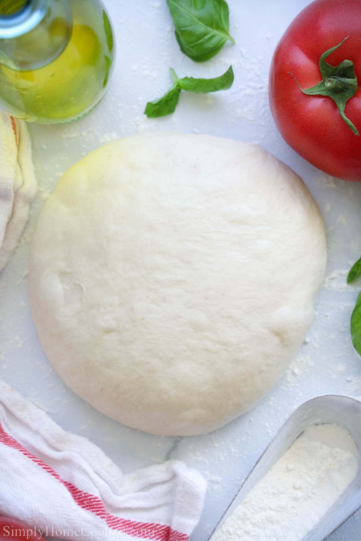 A simple and delicious pizza dough recipe that is made with minimal ingredients that you more likely already have at home now. #pizzadough #homemadepizza #pizzadoughrecipe #pizza #easypizzadough Homemade Pizza Dough with basil, tomato, and flour nearby.
