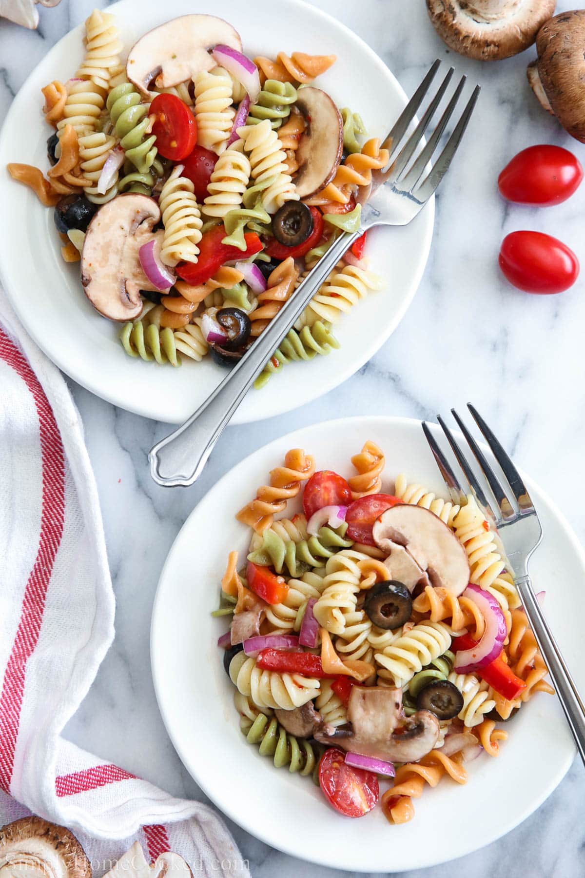 This Tri Color Pasta Salad is a very quick and easy way to add a colorful side to your lunch or dinner. It's so filling it could be considered a meal by itself. #pastasalad #italiansalad #tricolorpasta #easypastasalad #quicksalad Two plates of Tri Color Pasta Salad with forks and mushrooms and cherry tomatoes nearby.