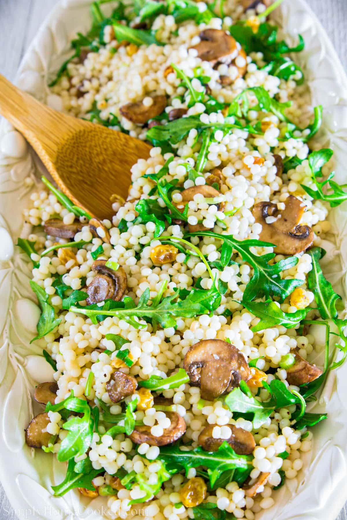This delicious couscous salad recipe is infused with white balsamic soaked raisins, crisp arugula, and sautéed garlic mushrooms. Light, healthy, and yummy! #couscous #couscoussalad #pastasalad #healthypasta #healthypastasalad Couscous Arugula Salad and a wooden spoon.