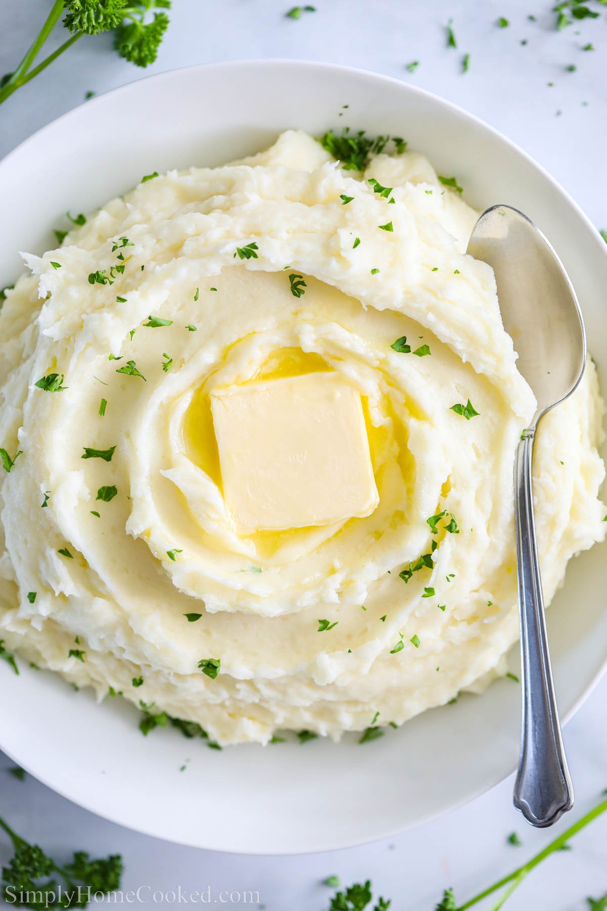 Bowl of Super Creamy Mashed Potatoes with a spoon and topped with a pat of butter and parsley.