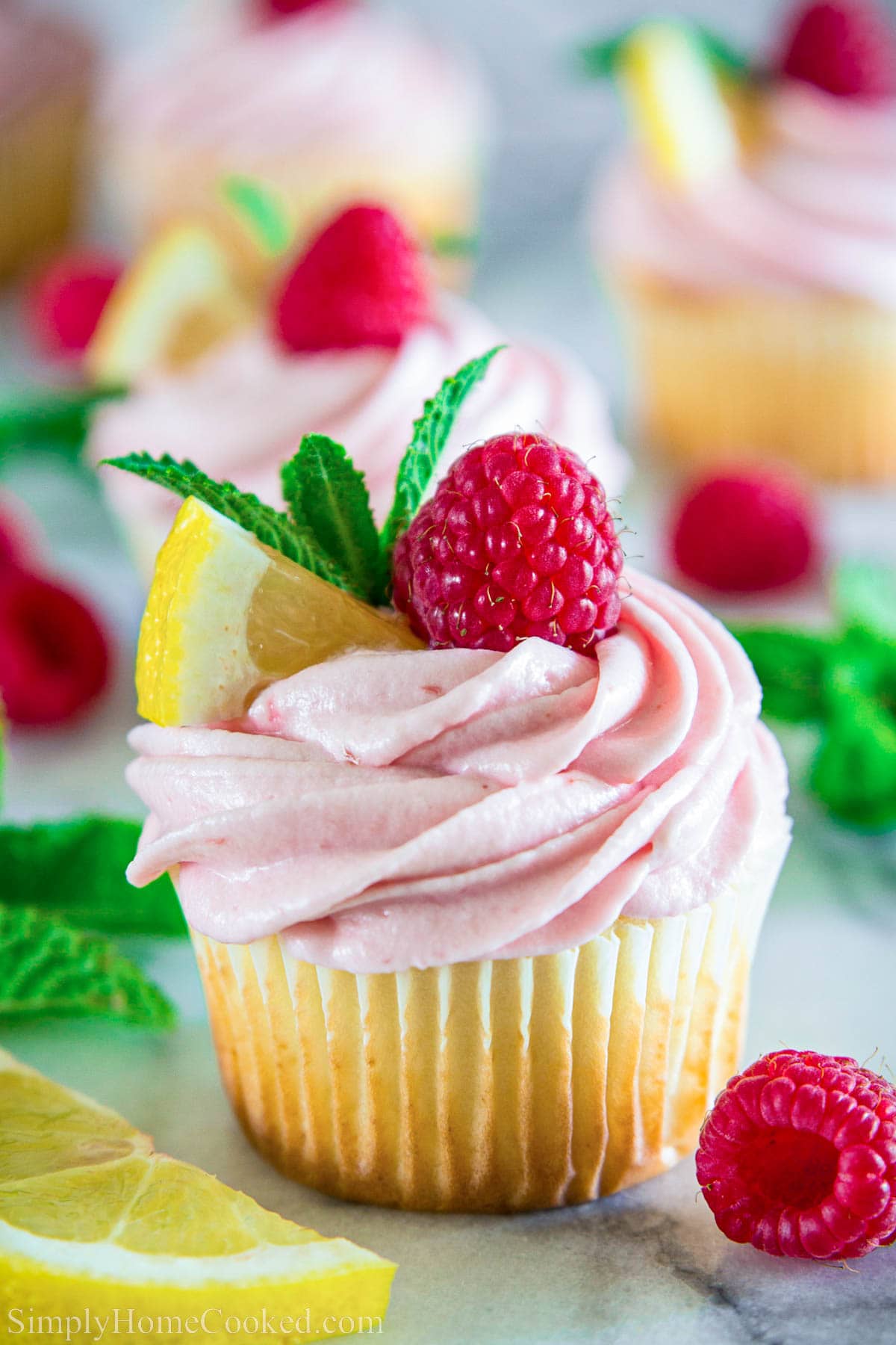 Lemon cupcakes with fresh raspberry buttercream. The ultimate summer dessert. #lemoncupcakes #raspberrybuttercream #buttercream #raspberrydessert #cupcakes Lemon Raspberry Cupcake topped with lemon, mint, and a raspberry with more nearby.